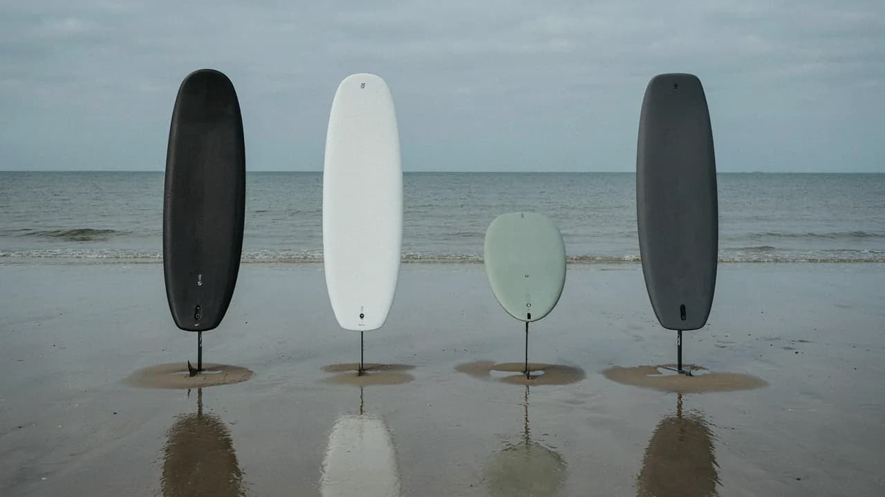 Quatre planches d’e-foil de formes et de finitions différentes, plantées côte à côte sur le sable au bord de l’eau